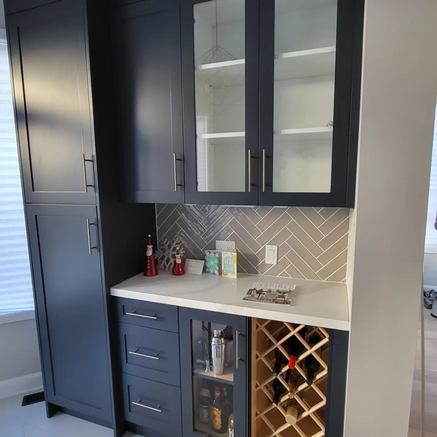 Navy blue built-in bar cabinet with glass doors, wine rack, herringbone tile backsplash, and white countertop